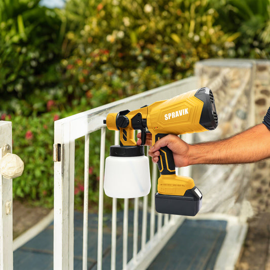 Person using a yellow and black Spravik paint sprayer on a white metal railing outdoors.