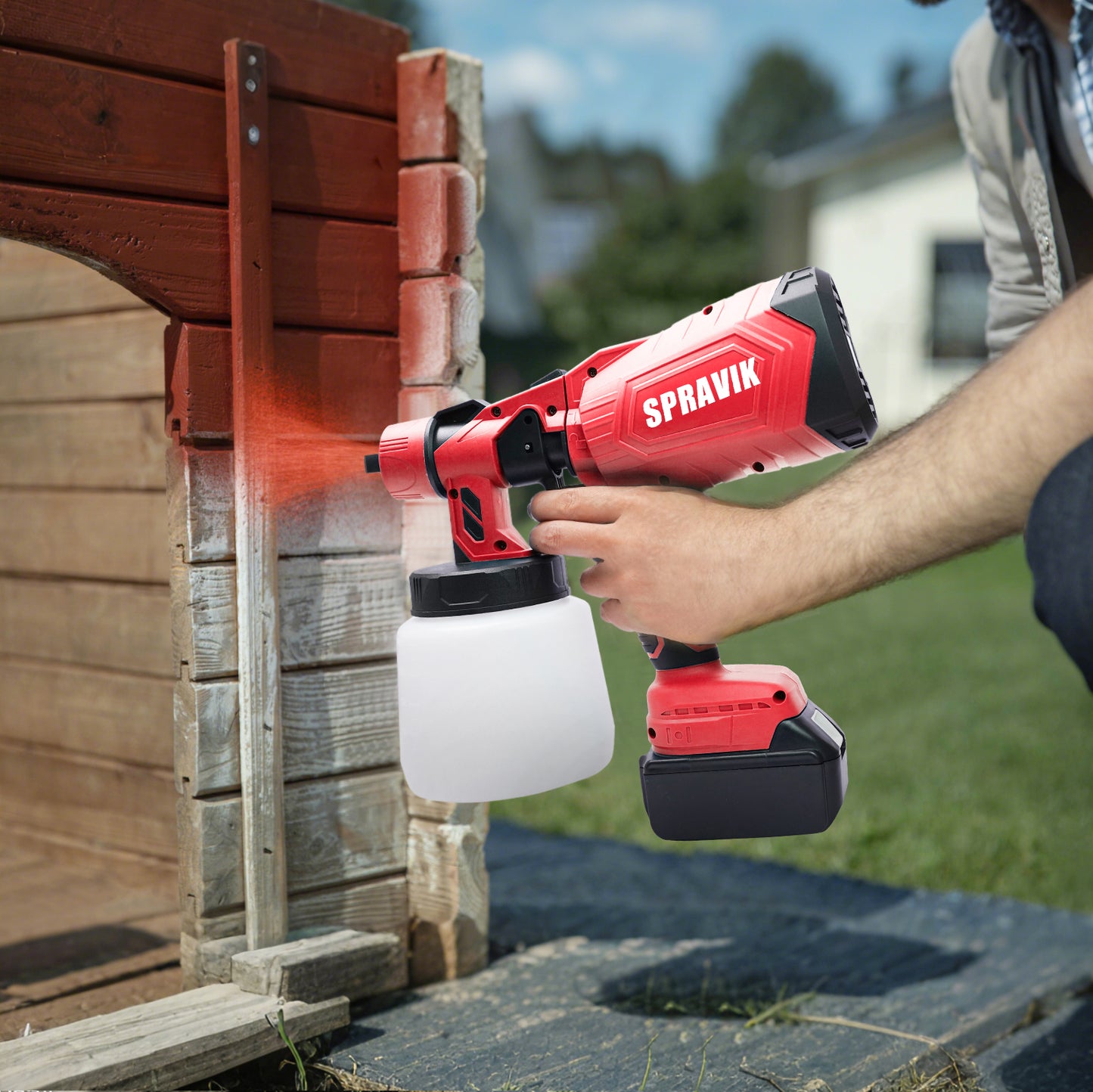 Person using a red Spravik paint sprayer to paint wooden planks outdoors.