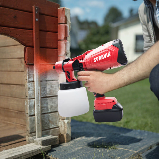Person using a red Spravik paint sprayer to paint wooden planks outdoors.